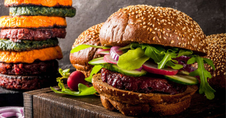 A vegan burger with raw vegetables on a whole wheat bun next to a pile of plant-based meat patties