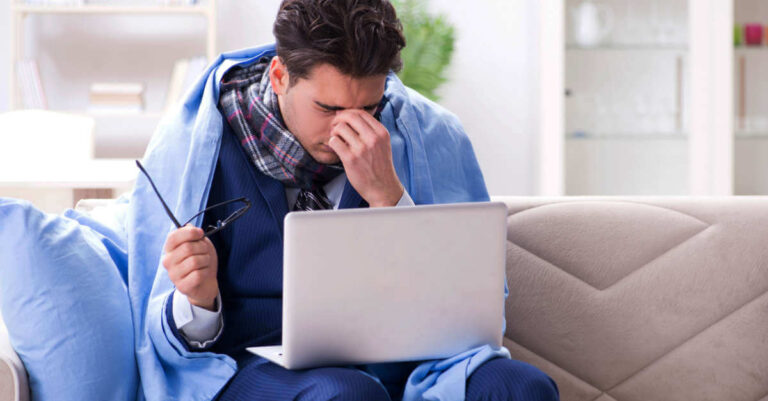 A male employee sitting on the couch trying to work on his laptop while feeling ill